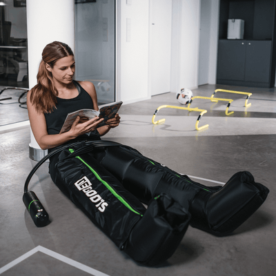 Sportlerin sitzt im Trainingsraum und liest ein Magazin, während sie Recovery Boots zur Muskelregeneration nach dem Workout trägt