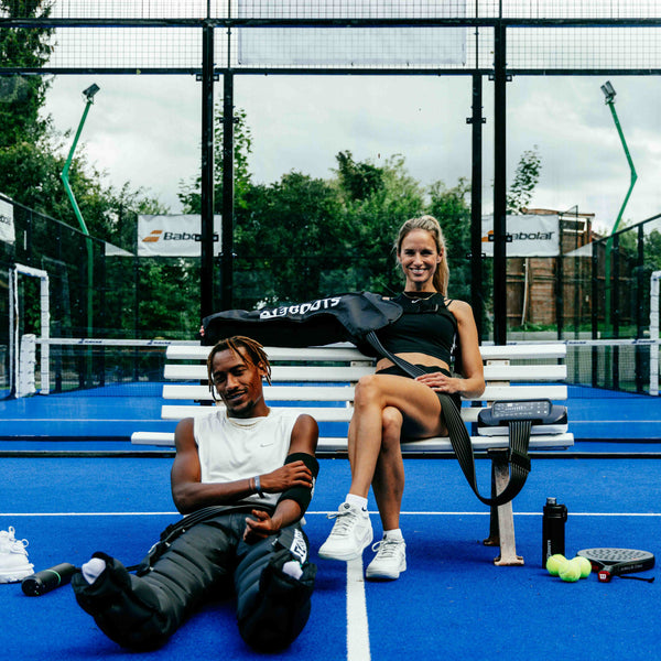 Reboots: Eine blonde Frau sitzt auf einer Bank neben einer Person mit dunkler Hautfarbe, die sich mit Sport-Drucktherapie erholt.