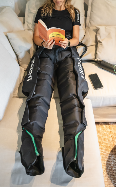 Sportlerin entspannt zu Hause auf dem Sofa, liest ein Buch und nutzt Recovery Boots zur Regeneration der Beinmuskulatur