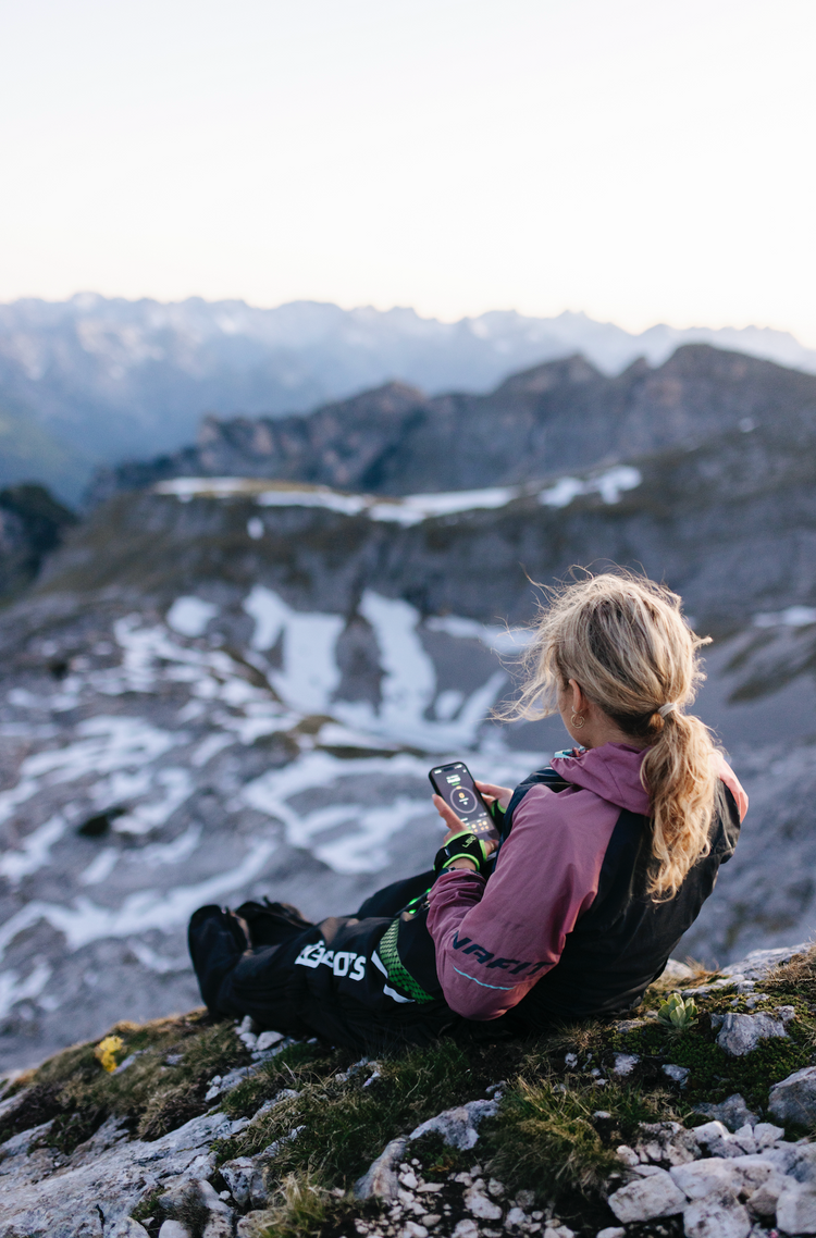 Sportlerin sitzt in den Bergen und nutzt Recovery Boots mit App-Steuerung zur Regeneration nach einer Bergtour
