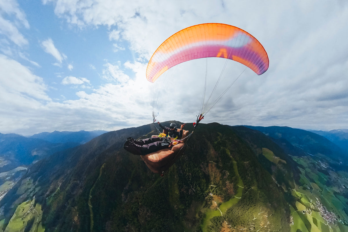 Paragleiter mit orangefarbenem Schirm fliegt über grüne Berge und Täler an einem sonnigen Tag