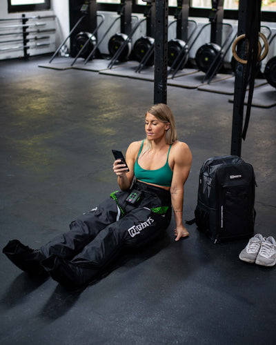 Athletin sitzt nach dem Training im Fitnessstudio auf dem Boden und nutzt Recovery Boots mit App-Steuerung zur Regeneration der Beine