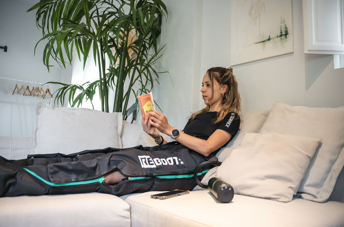 Frau entspannt auf dem Sofa, liest ein Buch und nutzt dabei die Reboots Recovery Boots zur Regeneration der Beine.
