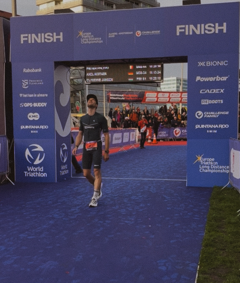 Triathlet überquert die Ziellinie bei einem Wettkampf, während Reboots als offizieller Recovery-Partner auf den Bannern sichtbar ist – Regeneration für Höchstleistungen im Ausdauersport.