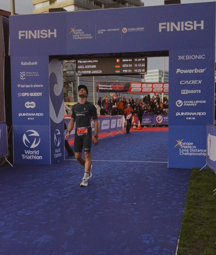 Triathlet überquert die Ziellinie bei einem Wettkampf, während Reboots als offizieller Recovery-Partner auf den Bannern sichtbar ist – Regeneration für Höchstleistungen im Ausdauersport.