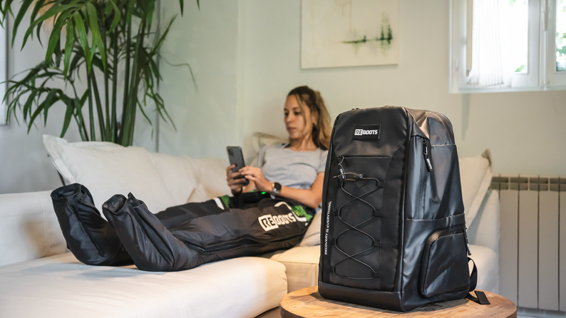 Athletin erholt sich mit Reboots Recovery Boots auf dem Sofa – im Vordergrund der stylishe, funktionale Reboots Rucksack für unterwegs.