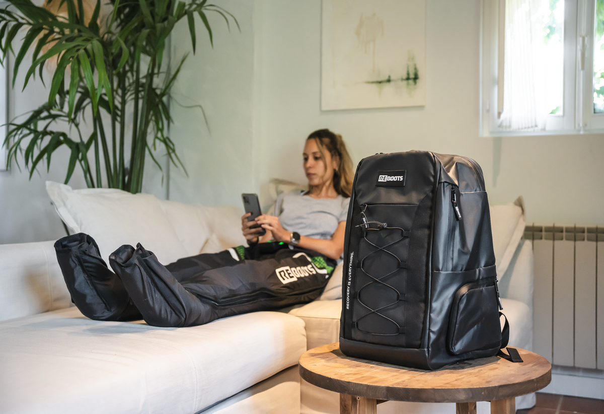 Athletin erholt sich mit Reboots Recovery Boots auf dem Sofa – im Vordergrund der stylishe, funktionale Reboots Rucksack für unterwegs.