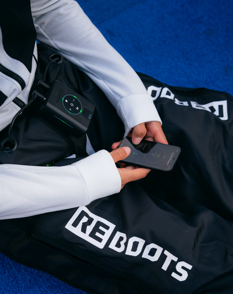 Athlet steuert Reboots Recovery Boots mit Smartphone – kabellose Kompressionsmassage für optimale Regeneration.
