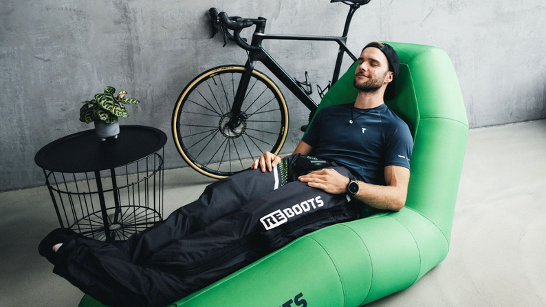Sportler entspannt auf einer Liege mit Reboots Recovery Boots zur Kompressionsmassage – Regeneration nach dem Radtraining im Wohnzimmer neben Rennrad.