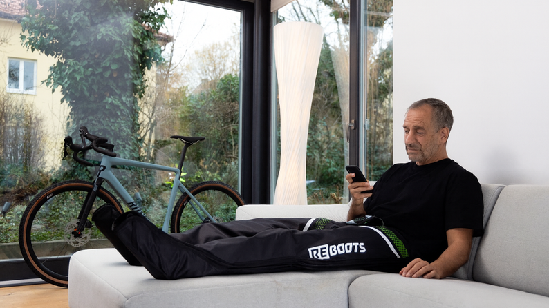 Mann entspannt auf Sofa mit Reboots Recovery Boots nach dem Radfahren – smarte Regeneration per App-Steuerung für bessere Durchblutung und schnellere Erholung der Beinmuskulatur.