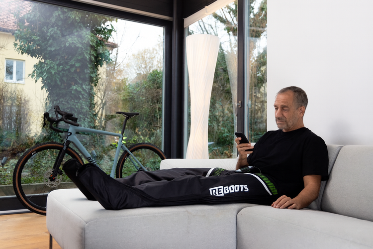 Mann entspannt auf Sofa mit Reboots Recovery Boots nach dem Radfahren – smarte Regeneration per App-Steuerung für bessere Durchblutung und schnellere Erholung der Beinmuskulatur.