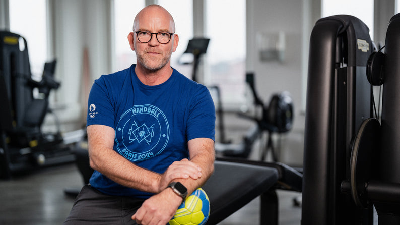 Dr. Philip Lübke, Facharzt für Orthopädie und Unfallchirurgie sowie Mannschaftsarzt des THW Kiel, sitzt in einem Fitnessstudio mit einem Handball in der Hand. Er trägt ein blaues Olympia Paris 2024 Handball-Shirt.