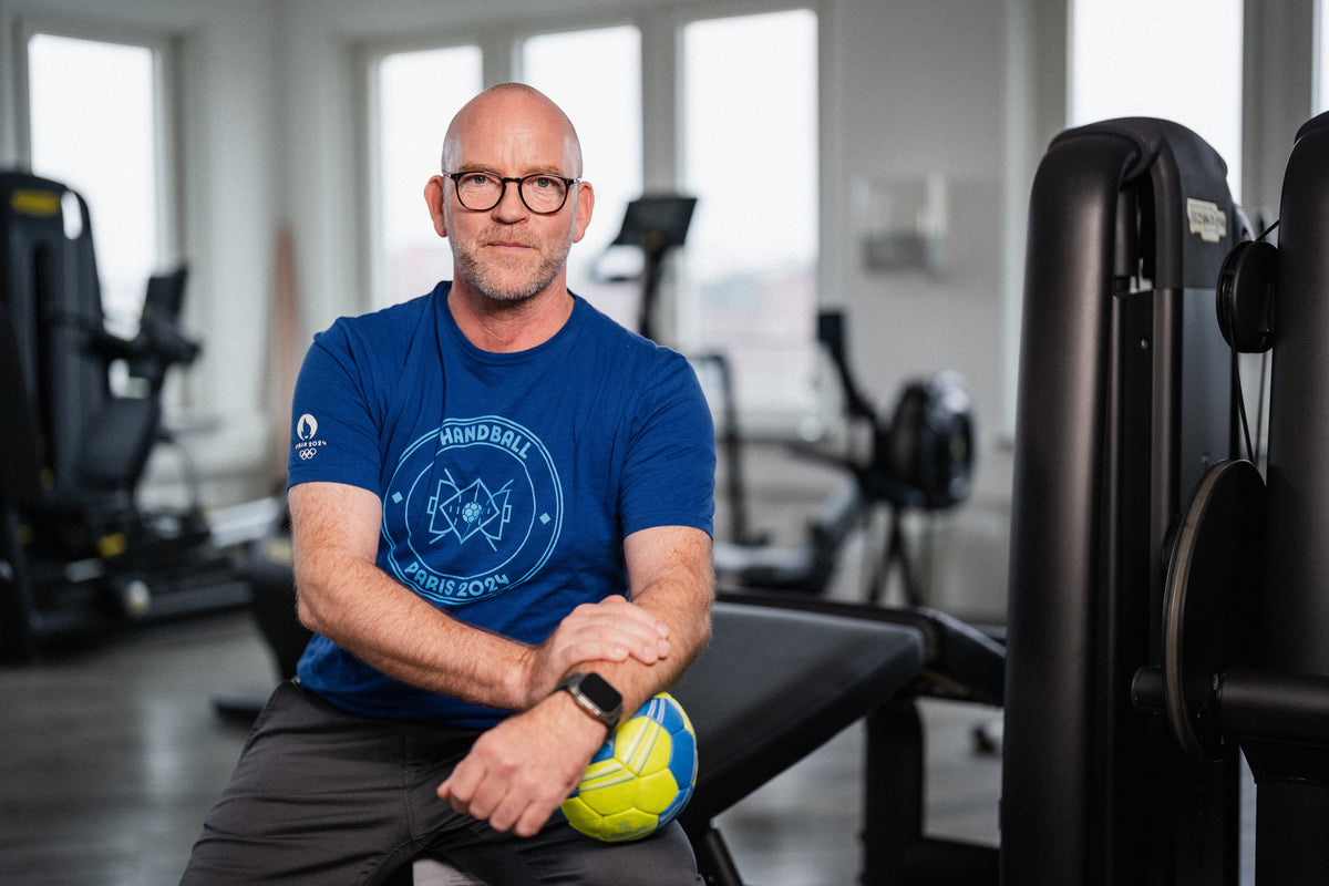 Dr. Philip Lübke, Facharzt für Orthopädie und Unfallchirurgie sowie Mannschaftsarzt des THW Kiel, sitzt in einem Fitnessstudio mit einem Handball in der Hand. Er trägt ein blaues Olympia Paris 2024 Handball-Shirt.