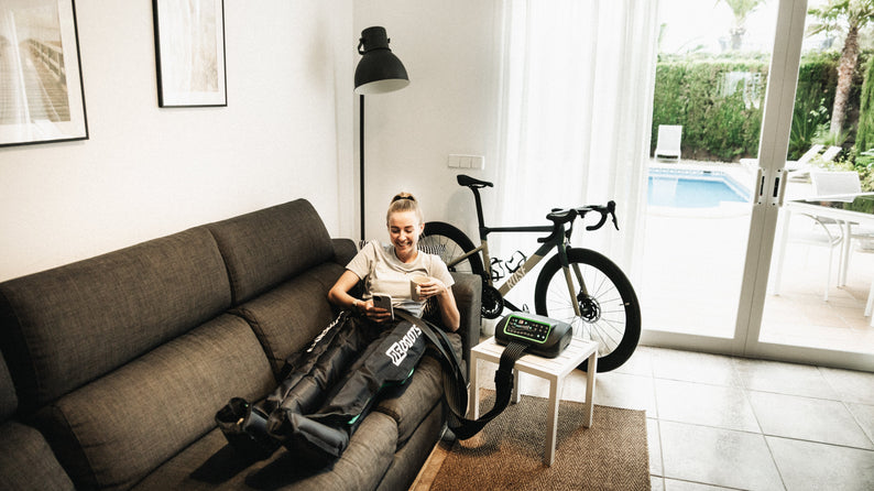 Sportlerin entspannt mit Reboots Recovery Boots auf dem Sofa – Kompressionsmassage zur Muskelregeneration nach dem Radtraining im Wohnzimmer.