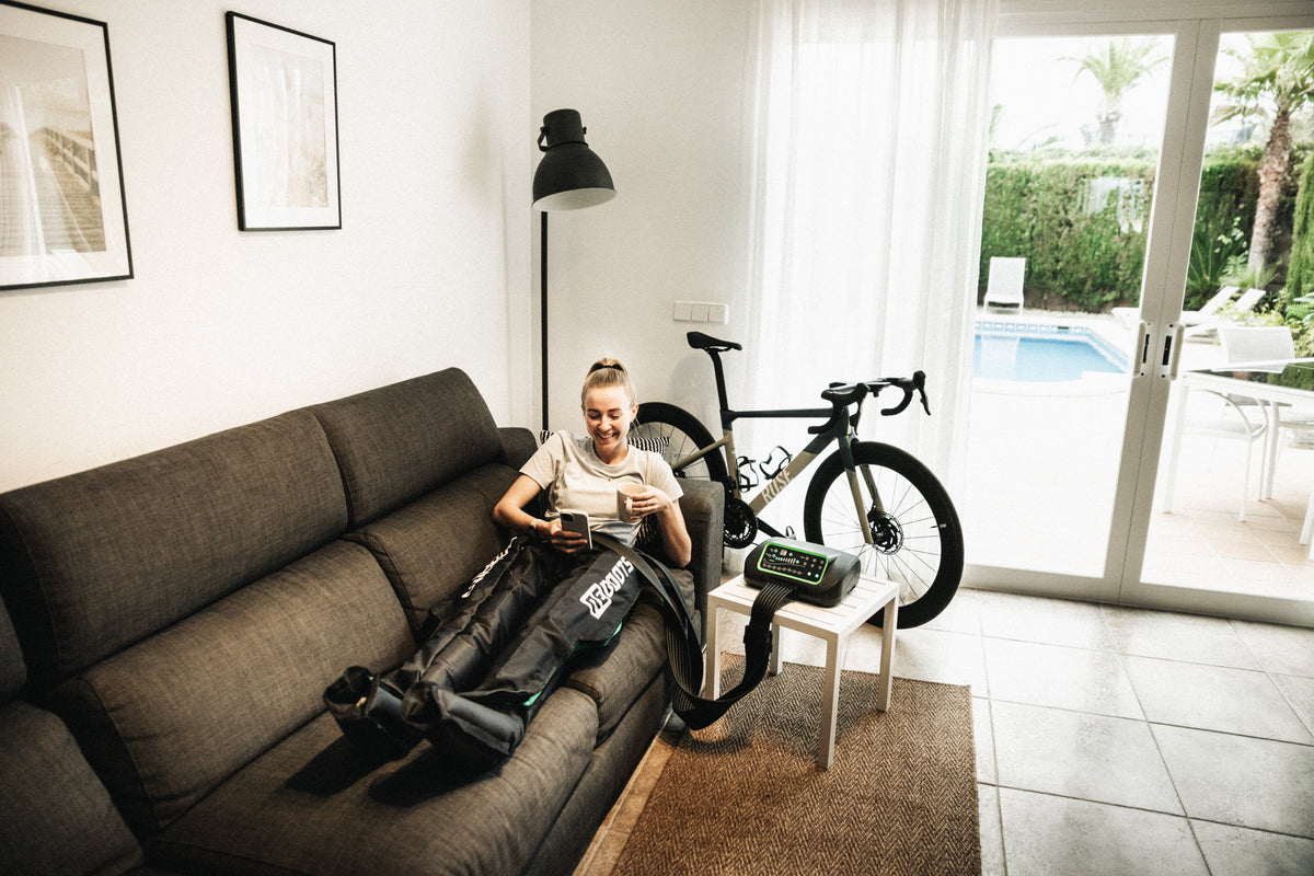 Sportlerin entspannt mit Reboots Recovery Boots auf dem Sofa – Kompressionsmassage zur Muskelregeneration nach dem Radtraining im Wohnzimmer.