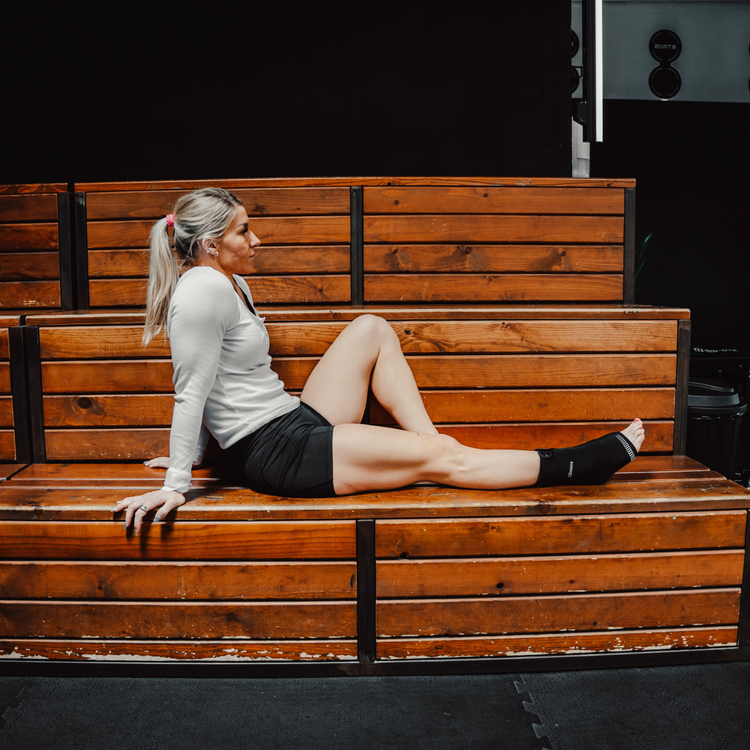 Sportlerin sitzt im Gym und nutzt Reboots Thermo Sleeve Ankle am Fuß zur Regeneration nach dem Workout.