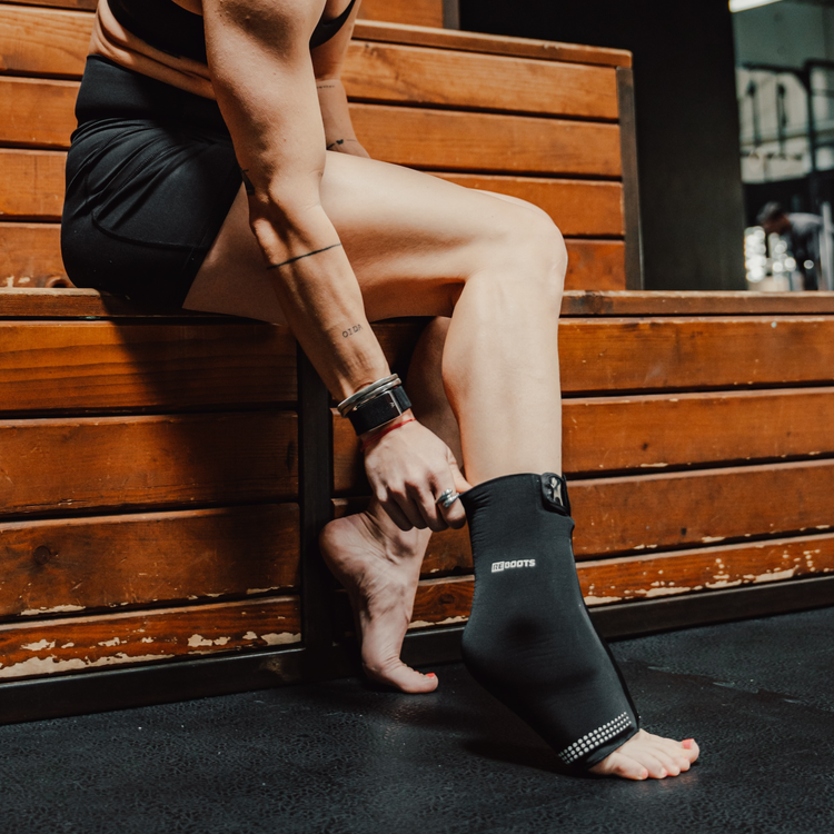 Sportlerin sitzt im Gym und nutzt Reboots Thermo Sleeve Ankle am Fuß zur Regeneration nach dem Workout.