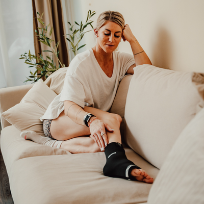 Sportlerin sitzt auf dem Sofa und nutzt Reboots Thermo Sleeve Ankle am Fuß zur Regeneration nach dem Workout.
