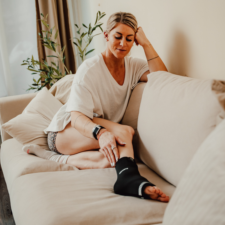 Sportlerin sitzt auf dem Sofa und nutzt Reboots Thermo Sleeve Ankle am Fuß zur Regeneration nach dem Workout.