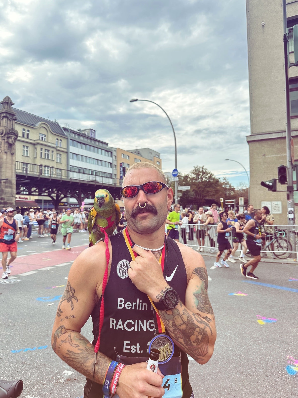 Rene Claußnitzer vom Reboots Athletes Project nach dem Berlin Marathon, lächelnd mit Papagei auf der Schulter und Medaille um den Hals im Trubel des Zieleinlaufs.