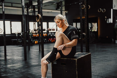 Sportlerin sitzt im Gym und nutzt Reboots Thermo Sleeves an Schulter und Fuß zur Regeneration nach dem Workout.