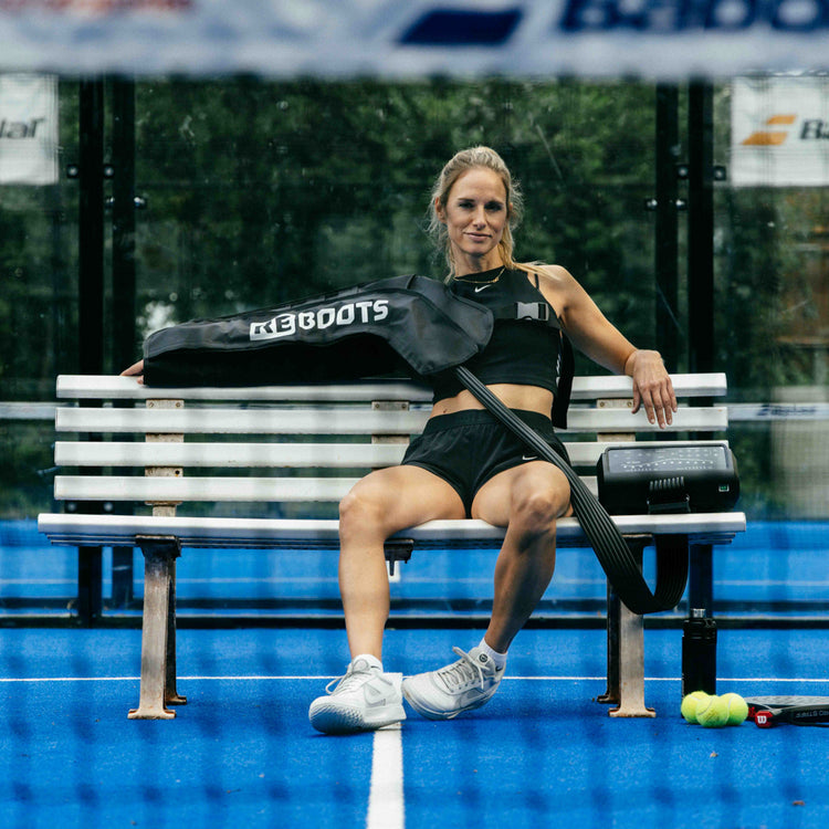 Reboots: Mujer rubia con ropa negra sentada en un banco, regenerando sus brazos con la terapia de presión deportiva One PRO.