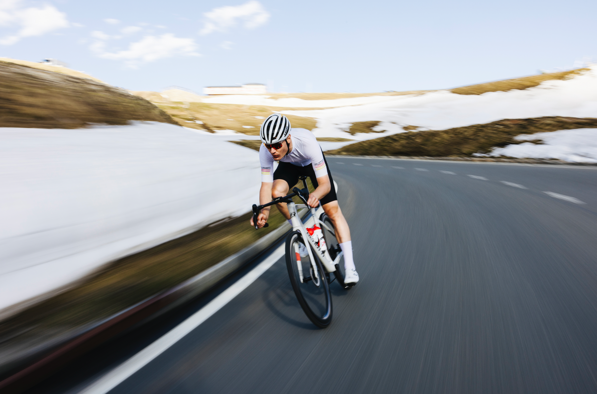 Radfahrer im weißen Trikot fährt mit hoher Geschwindigkeit eine Bergstraße mit Schneeresten in den Alpen hinunter