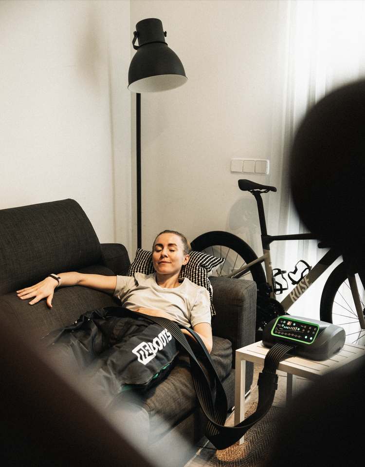 Radsportlerin erholt sich zu Hause mit den Reboots Recovery Boots auf dem Sofa – effektive Kompressionsmassage zur Regeneration der Beine nach dem Training.