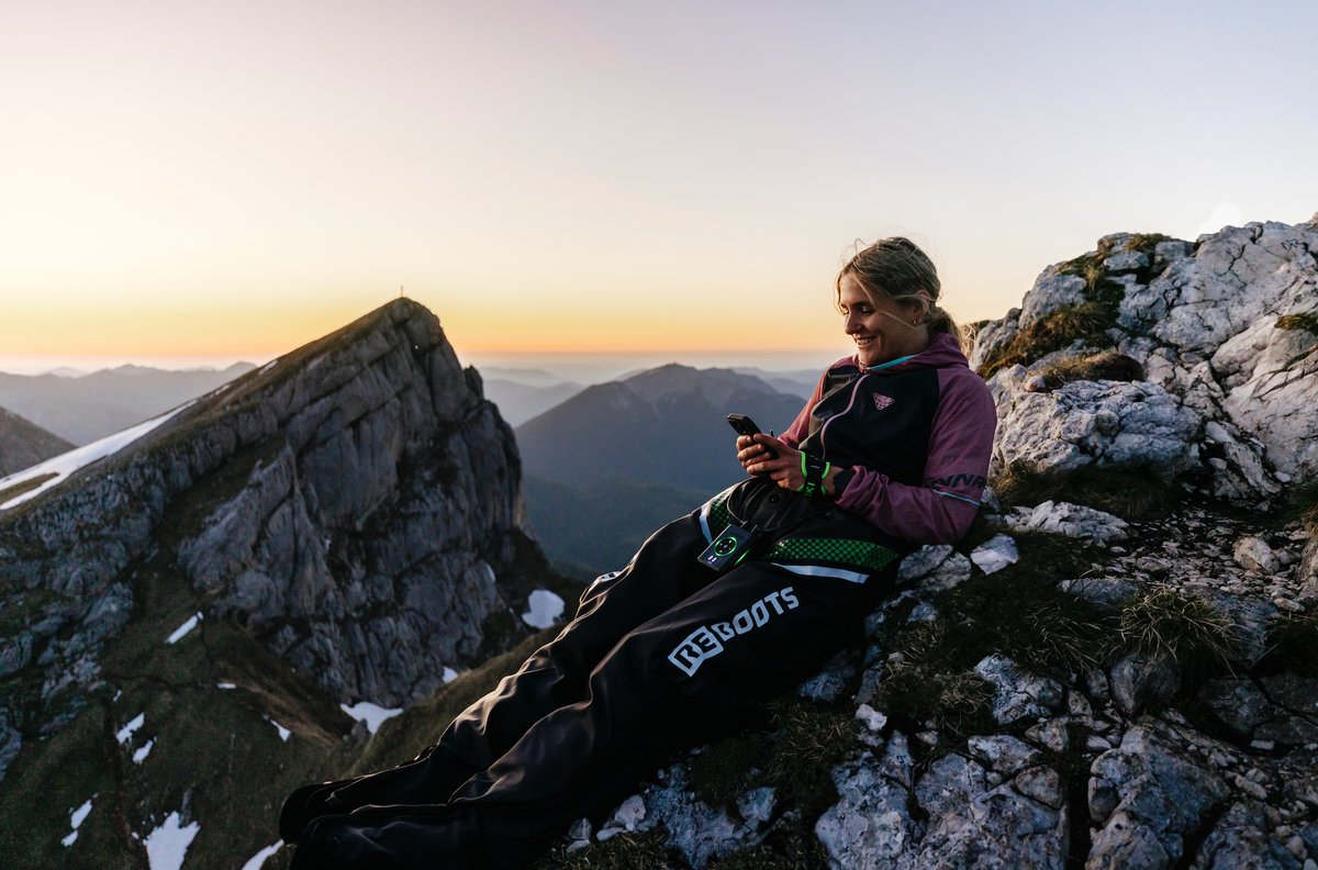 Athletin erholt sich mit Reboots Recovery Boots in den Bergen bei Sonnenuntergang – mobile Regeneration nach dem Sport mit beeindruckendem Alpenpanorama.