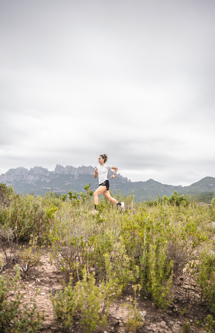 Trailrunnerin läuft dynamisch durch bergige Naturkulisse – Outdoor-Lauftraining vor felsigen Bergen unter bewölktem Himmel.
