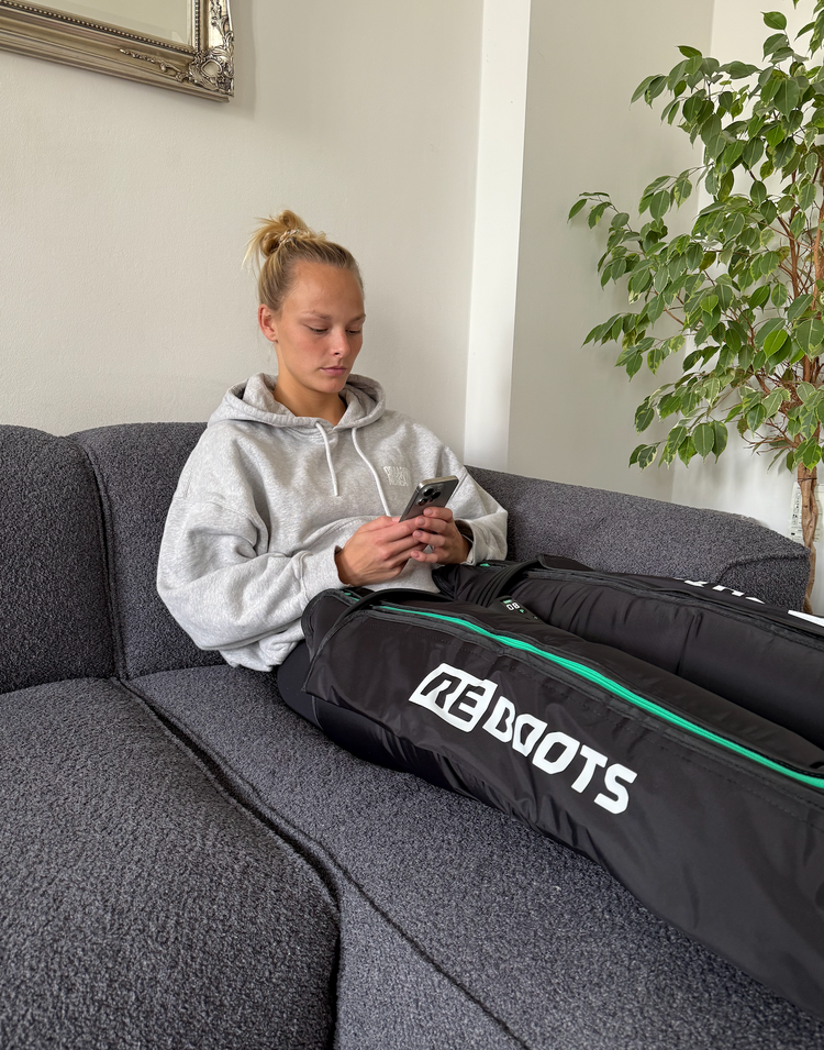 Ironman Athletin erholt sich zu Hause auf dem Sofa mit Reboots Recovery Boots und nutzt ihr Smartphone – effektive Regeneration nach intensivem Training.“