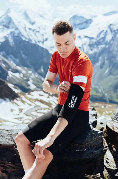 Radsportler legt in den Bergen eine Pause ein und zieht den Reboots Thermo Sleeve zur Armregeneration an, umgeben von verschneiten Alpen.