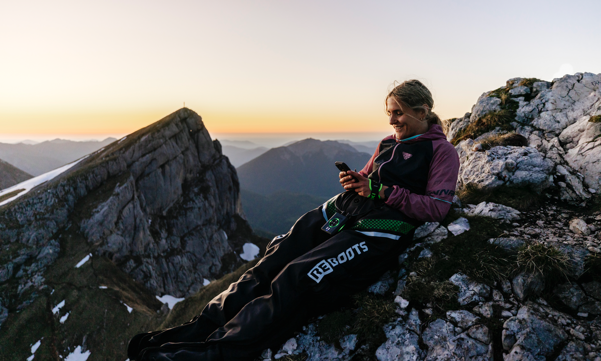 Athletin erholt sich mit Reboots Recovery Boots in den Bergen bei Sonnenuntergang – mobile Regeneration nach dem Sport mit beeindruckendem Alpenpanorama.