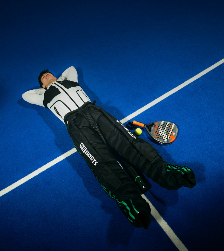 Padelspieler entspannt nach dem Match auf dem Court und nutzt Reboots Recovery Pants zur schnellen Beinregeneration durch Kompressionsmassage.“