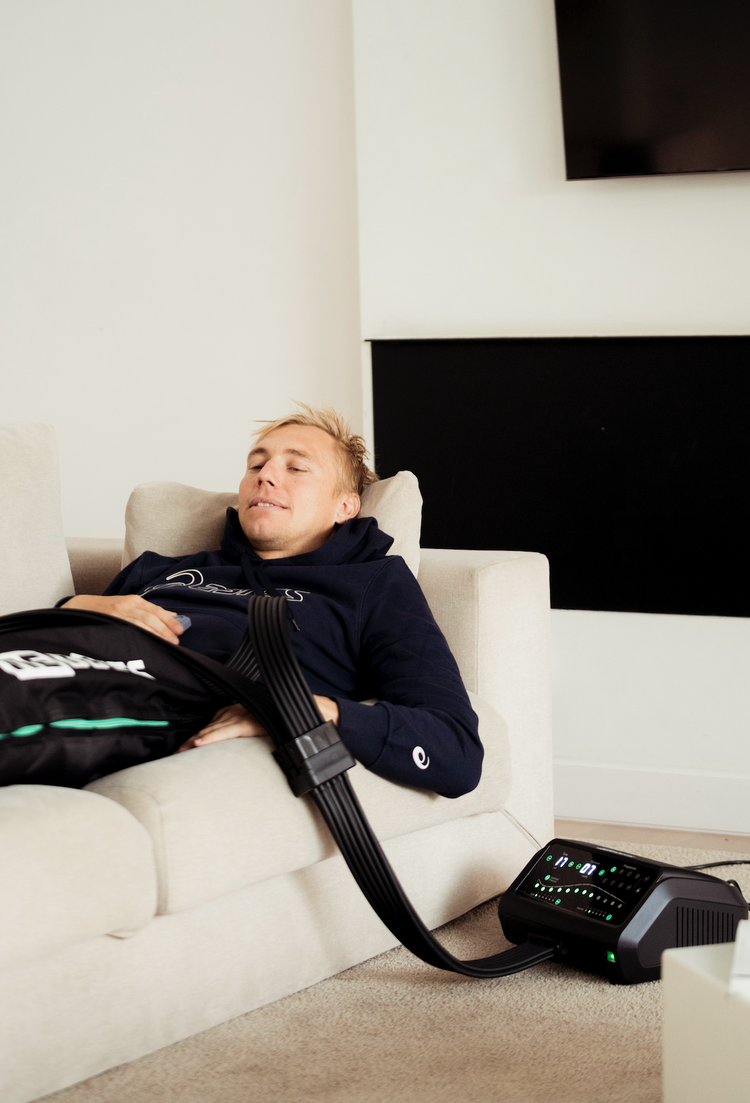Triathlet regeneriert zu Hause auf dem Sofa mit Reboots Kompressionsmassage für Beine nach intensivem Ausdauertraining.
