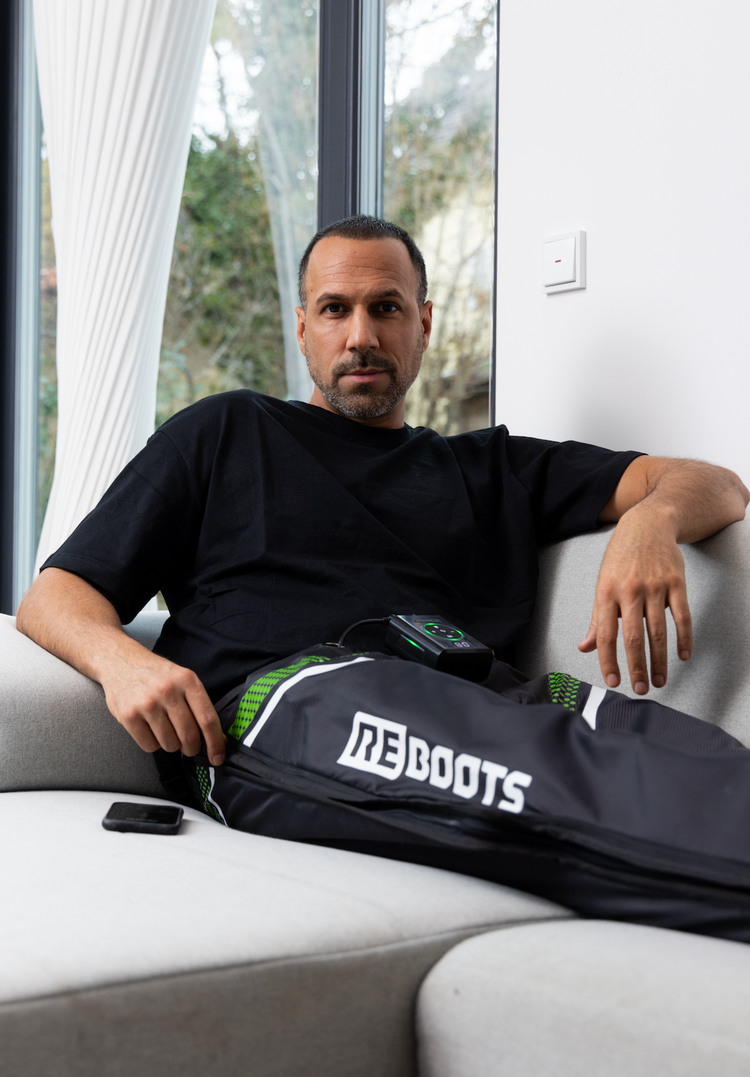 Mann entspannt auf dem Sofa mit Reboots Recovery Boots – kabellose Kompressionsmassage zur Muskelregeneration und schnelleren Erholung nach dem Sport.