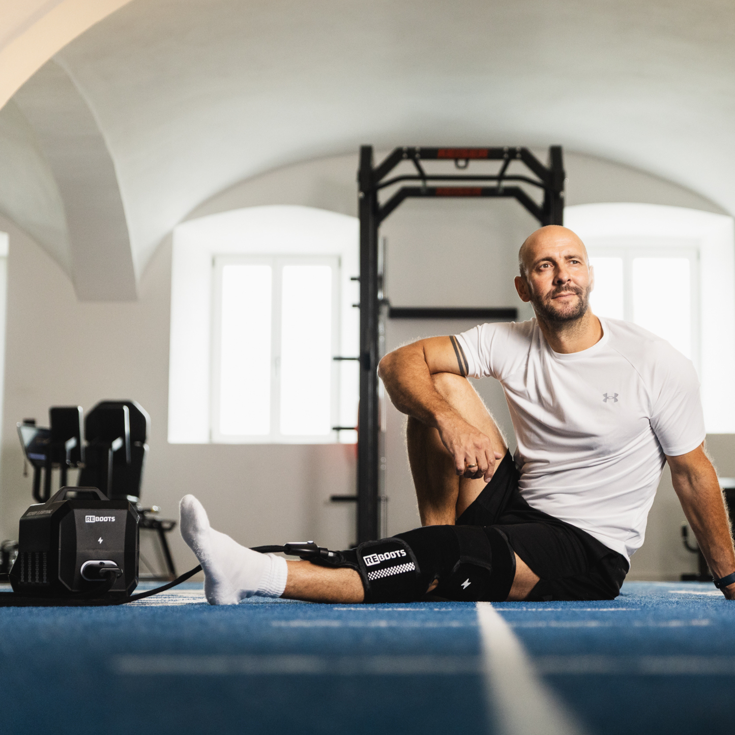 Hombre sentado en el suelo del gimnasio usando el Reboots Cryo One con una rodillera para terapia de frío y compresión durante la regeneración.
