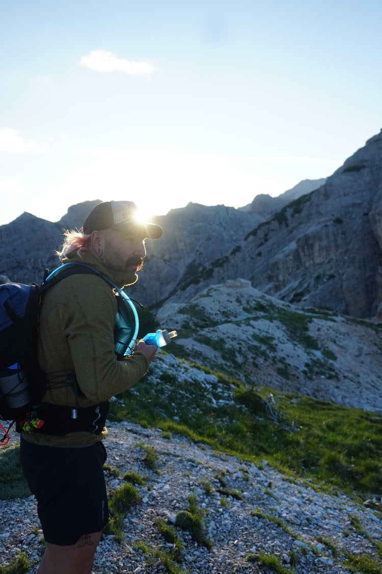 Mann mit Bart steht in den bergen. Er ist ein Hiker. Er hat eine kurze Hose, eine Windjacke und einen Hiker Rucksack an. Außerdem hält er eine Trinkflasche in der hand und guckt in die Ferne. 