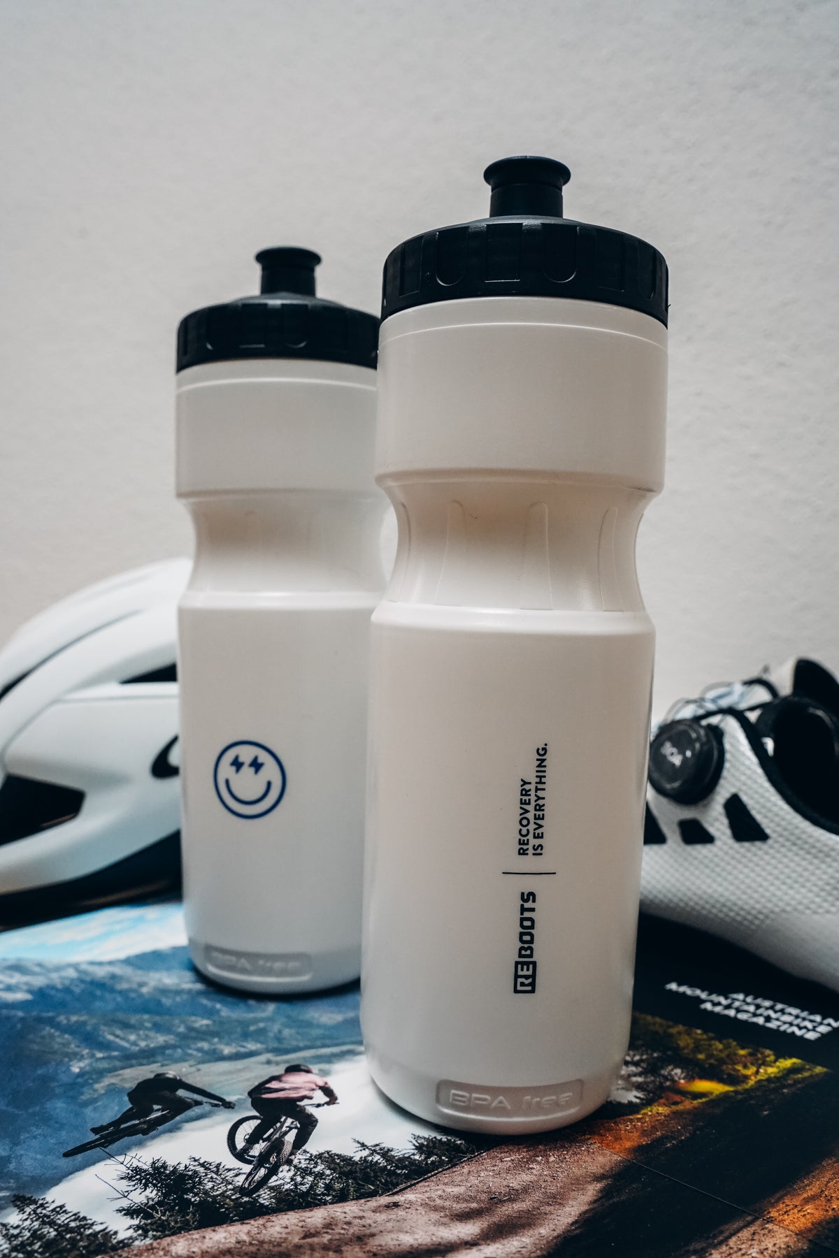Dos botellas deportivas blancas Reboots con el estampado "Recovery-Is-Everything", fotografiadas junto a un casco de ciclismo y zapatillas de ciclismo – Accesorios de ciclismo Move & Recover de Reboots