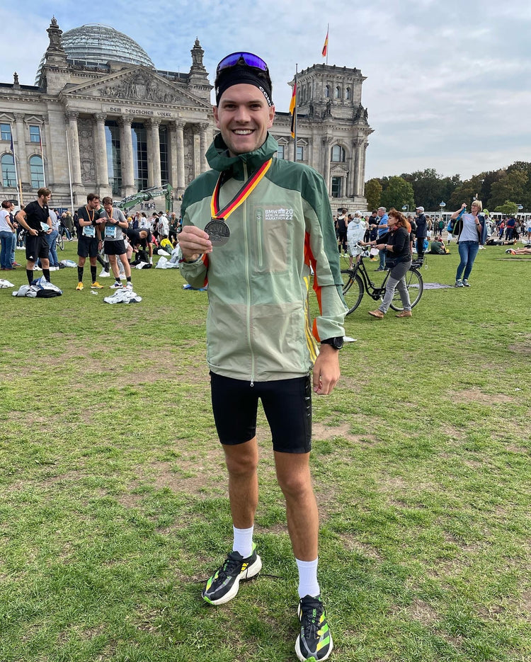 Ein junger Sportler in Sportklamotten steht or einem großen Gebäude mit einer Medaille um den Hals. Diese hält er lachend in die Kamera. 