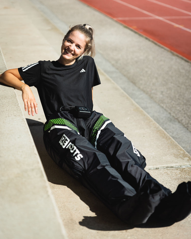 Sportlerin entspannt mit Reboots Recovery Boots auf der Laufbahn – mobile Kompressionsmassage zur schnelleren Beinregeneration nach dem Training.