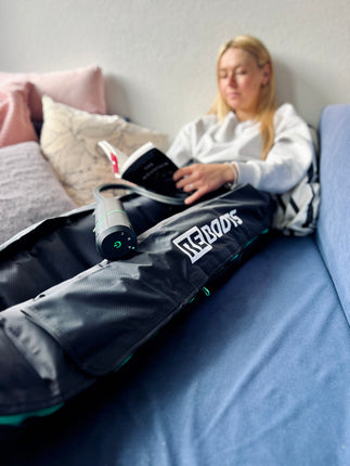 Reboots: Frau auf Sofa entspannt mit Buch und Beinkompressionsmassage