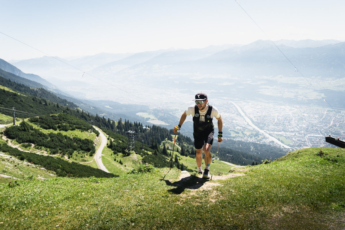 Ein junger Mann wandert den Berg hoch. Es sieht nach Trailunning aus. Er hat eine kurze Sporthose, ein helles t Shirt und eine Laufweste an. Außerdem trägt er Cap und Sonnenbrille. In de Händen hält er Walking Sticks. 