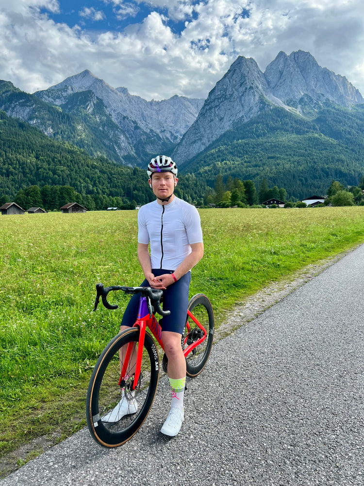 Ein junger, männlicher Rennradfahrer sitzt auf seinem roten Rennrad vor einer Bergkulisse. Füße sind auf dem Boden. Er trägt Fahrradtrikot, Fahrradhose, Helm und hat Kopfhörer im Ohr. 