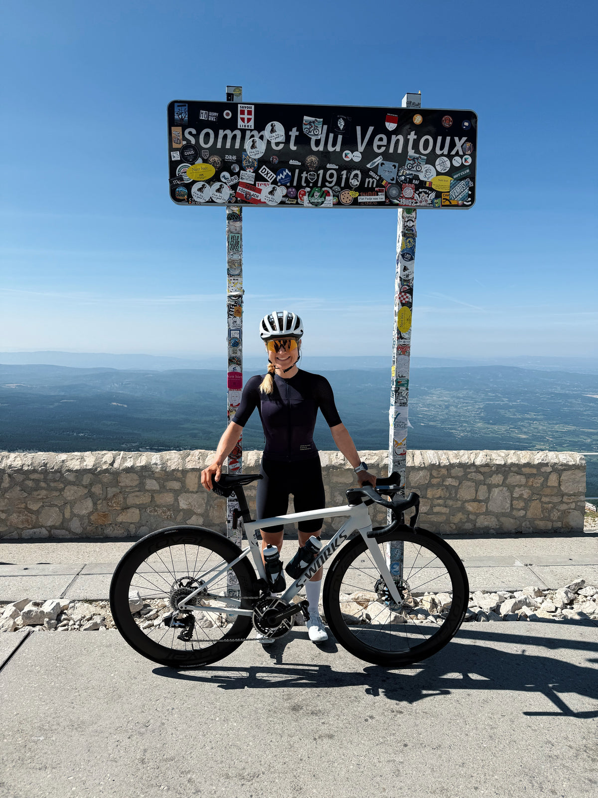 Frau steht auf dem Berg vor einem Schuld mit ihrem Rennrad in der Hand. 