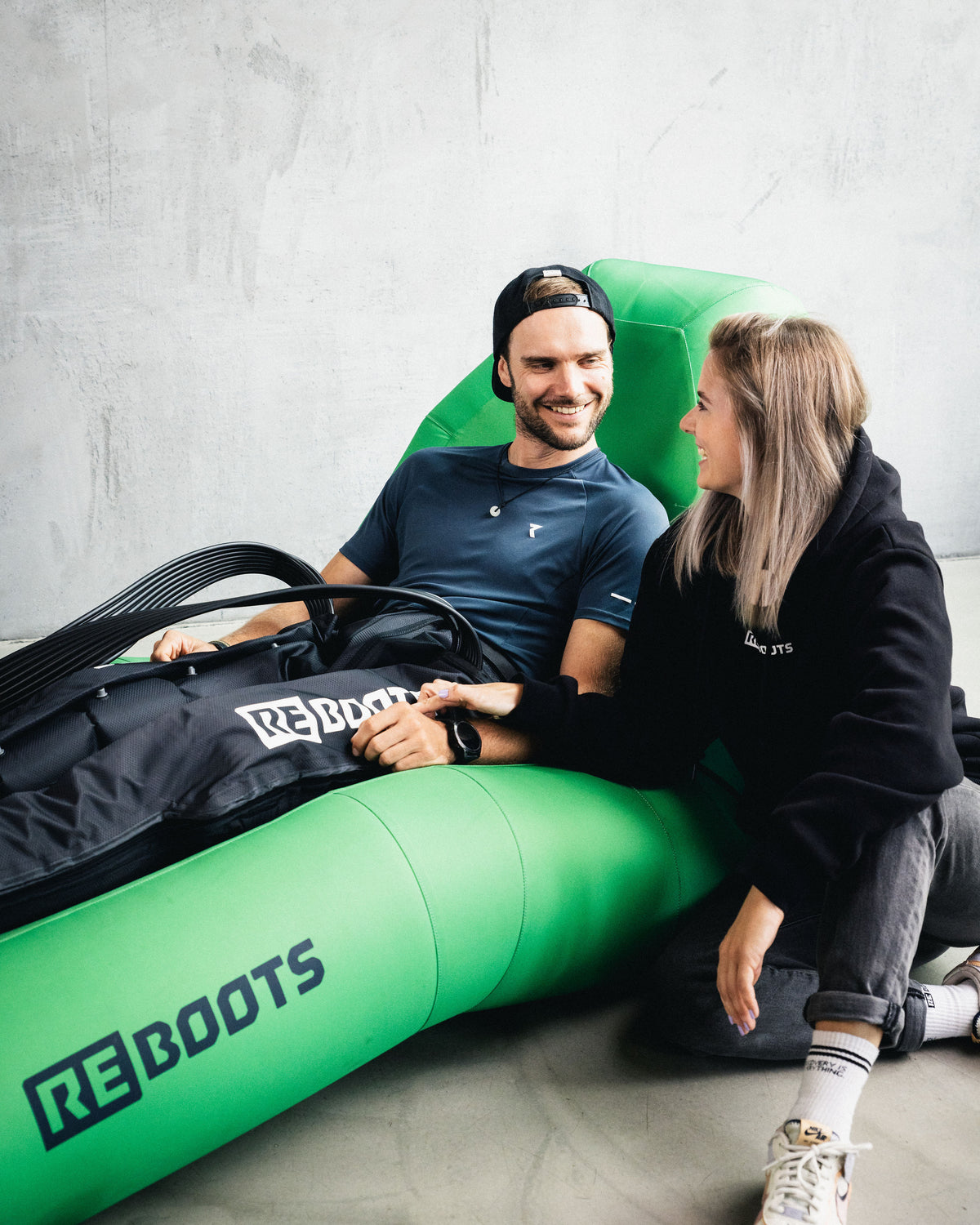 Sportler nutzt Reboots Recovery Boots im grünen Relax-Sessel – Kompressionsmassage zur Muskelregeneration, begleitet von Partnerin im Indoor-Setup.