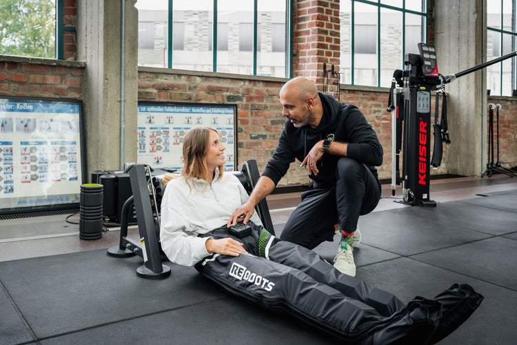 Sportlerin nutzt ein mobiles Reboots Kompressionsmassagegerät zur Regeneration der Beine im Fitnessstudio, während ein Trainer die Anwendung begleitet.