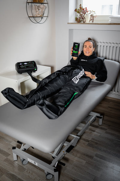 Frau liegt in Reboots One Pro Recovery Boots auf einer Therapieliege und steuert die Anwendung per Smartphone-App.