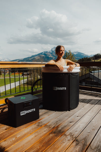Frau genießt eine Kälteanwendung im Reboots Ice Tub mit Chiller auf einer Sonnenterrasse mit Bergblick – Kältetherapie zur Regeneration und Erholung.