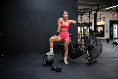 Sportlerin in pinkem Sportoutfit posiert nach dem Training im Fitnessstudio neben einem Rogue Airbike, Medizinball und Kurzhantel
